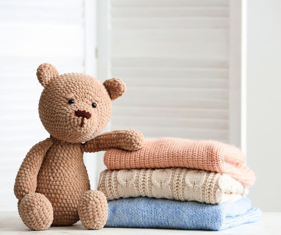 A crochet teddy bear leaning on a stack of pastel-colored knitted blankets in a softly lit room.