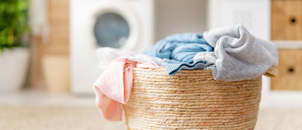 A wicker laundry basket filled with clothes, including jeans and shirts, sits in a bright room with a washing machine in the background.