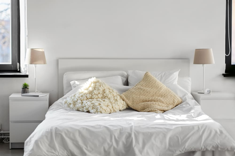 A bright bedroom with a neatly made white bed, flanked by two nightstands with lamps, and three textured pillows on the bed.