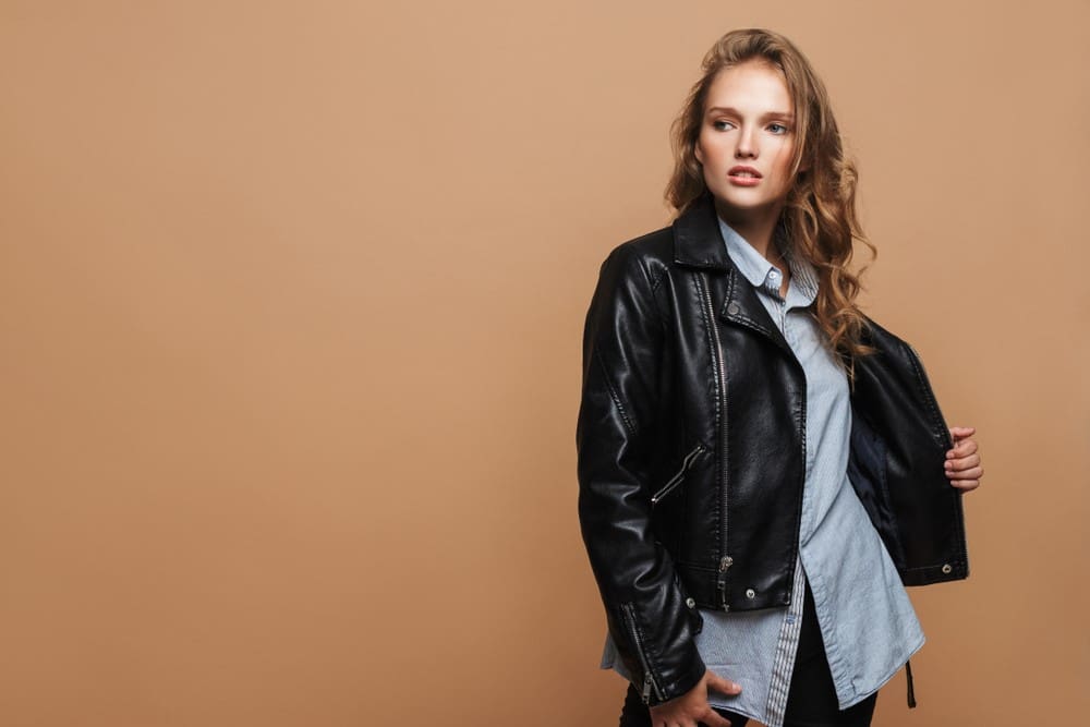 A young woman in a stylish leather jacket and denim shirt poses against a beige background.