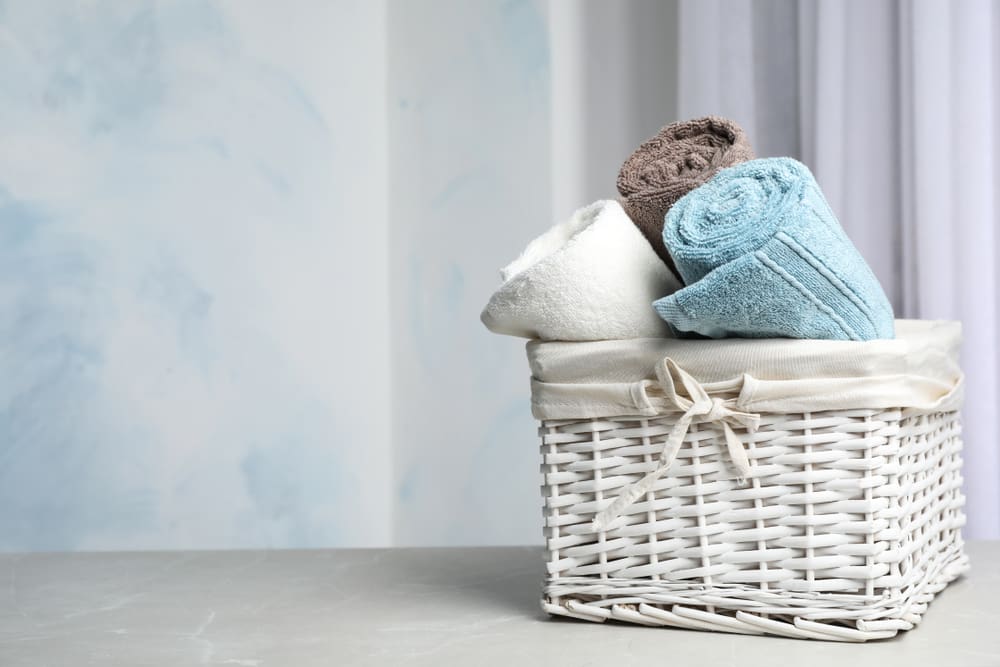 A white wicker basket containing neatly folded towels in white, brown, and blue, placed on a marble surface against a light blue background.