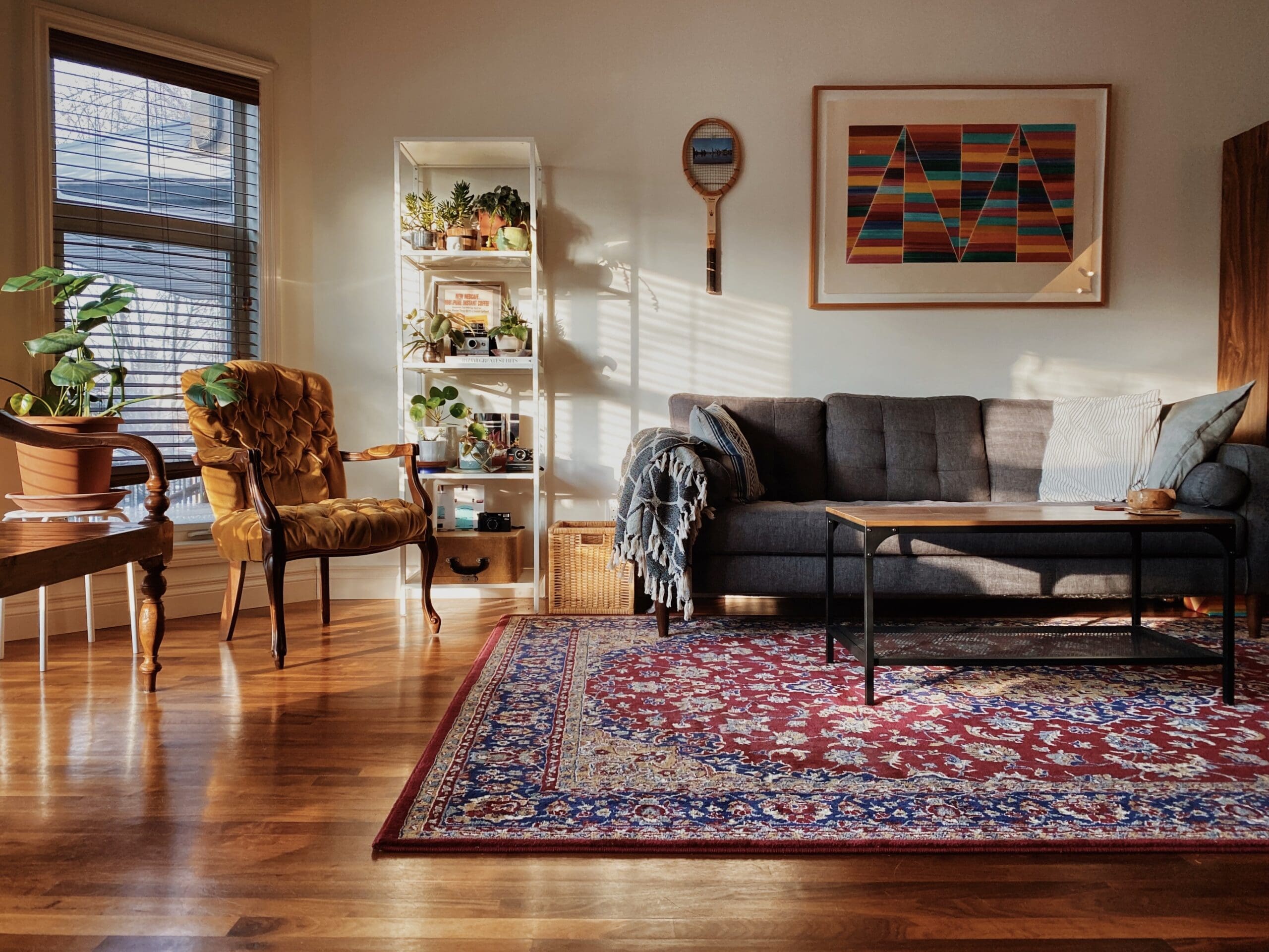 Sunlit living room featuring a sofa, armchair, coffee table, bookshelf, and colorful rug, with a decorative wall art and warm wooden floors.