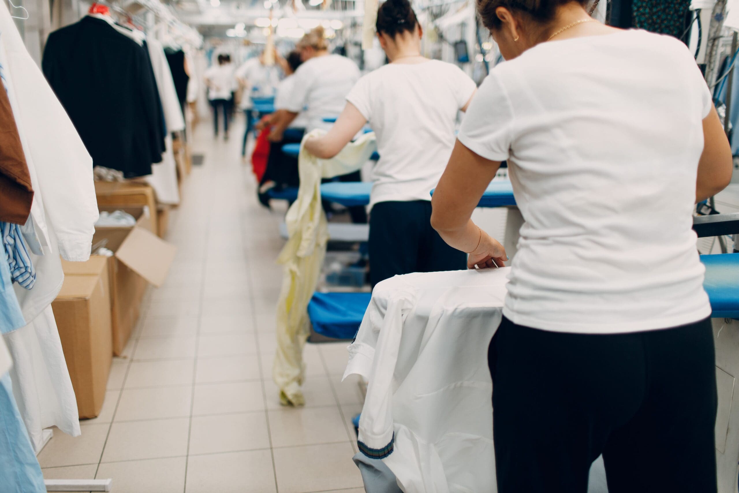 Workers in a laundry facility are seen ironing clothes. The scene is busy, with multiple people in white shirts focused on their tasks.