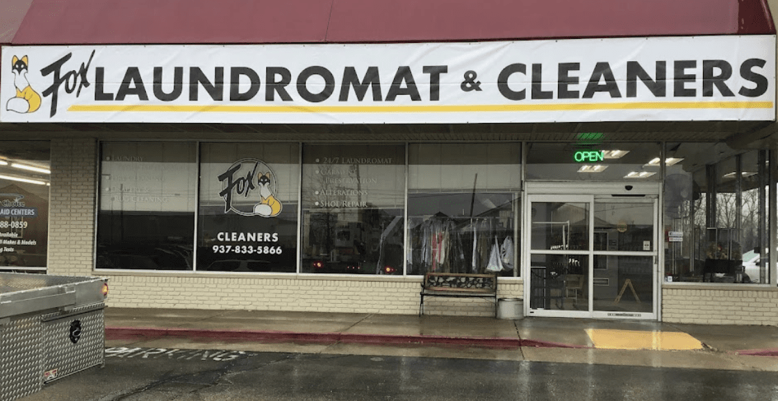 Street view of Fox Laundromat & Cleaners, featuring a glass storefront with an open sign, hanger racks inside, and a large white sign with a fox logo above the entrance.
