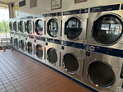 A row of ten front-loading industrial dryers in a laundromat with tiled floors and large windows.