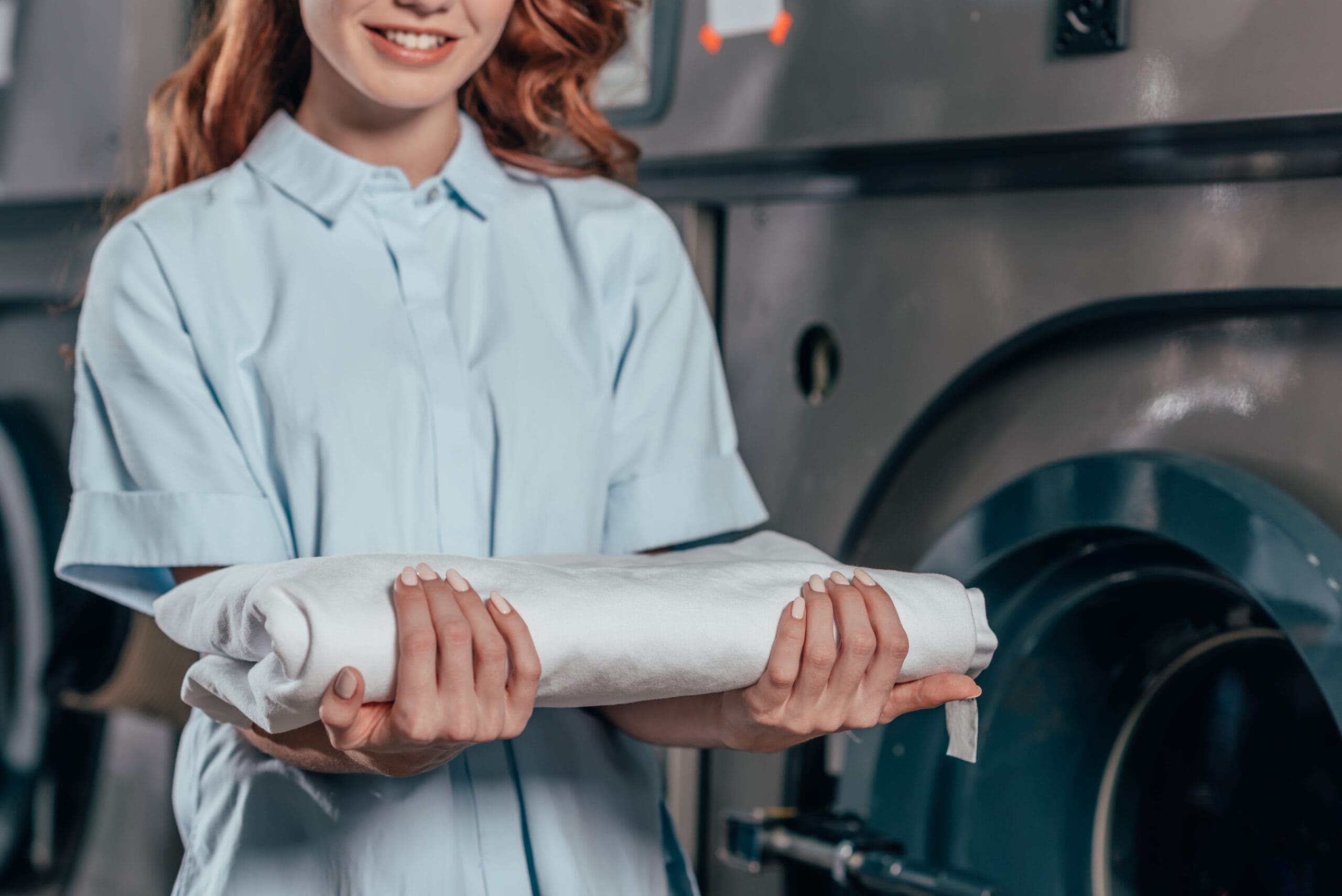 A person in a light blue shirt holds a neatly folded white cloth in front of a large industrial washing machine at the premier Oxford Dry Cleaner.