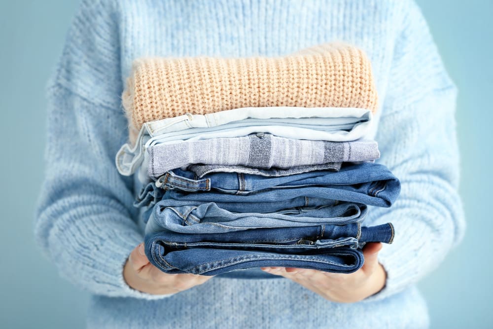 Person in a blue sweater holding a stack of folded clothing, including sweaters and jeans, against a light blue background.