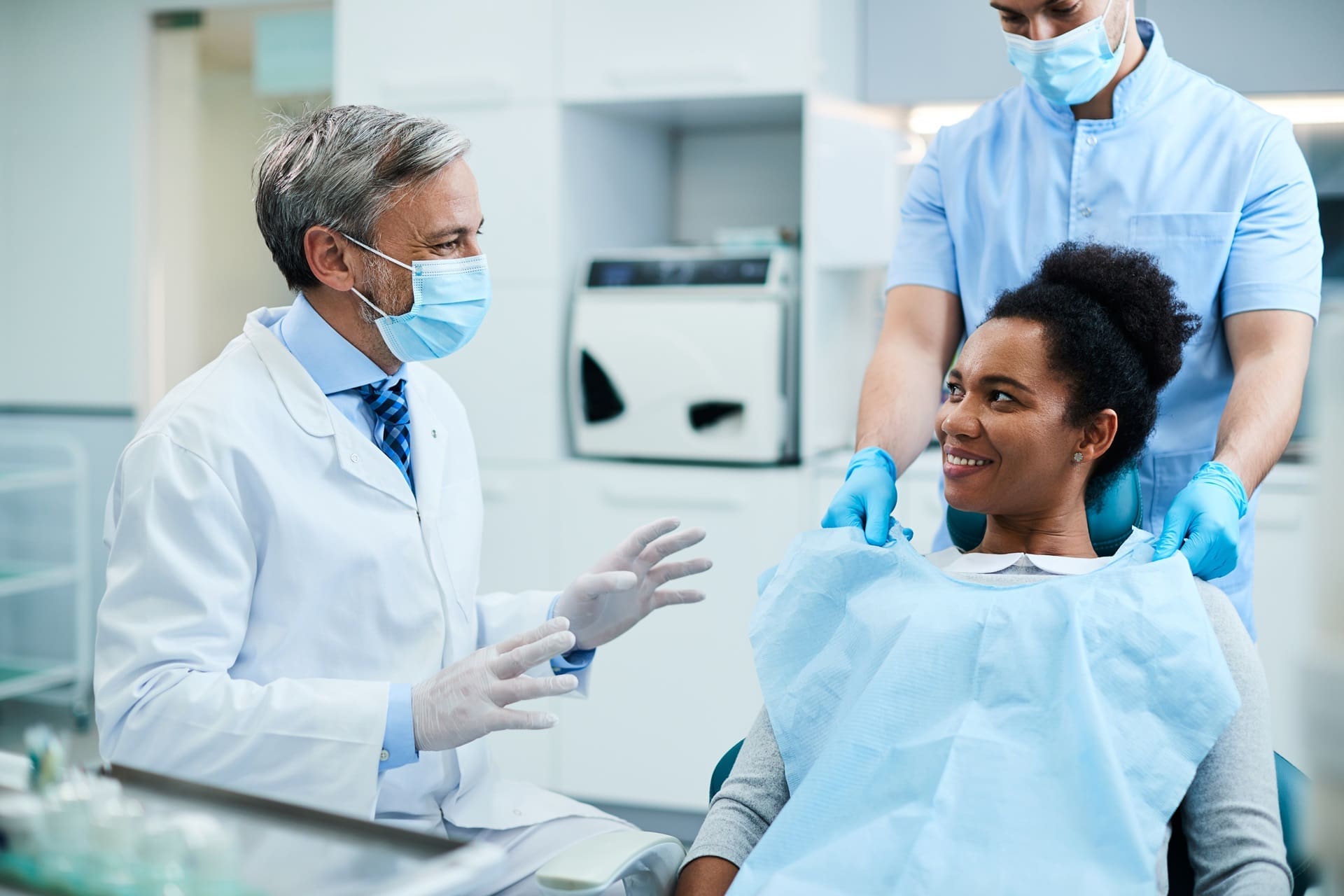 A dentist talks to a smiling patient sitting in a dental chair, while an assistant stands nearby. Both the dentist and assistant are wearing masks and gloves.