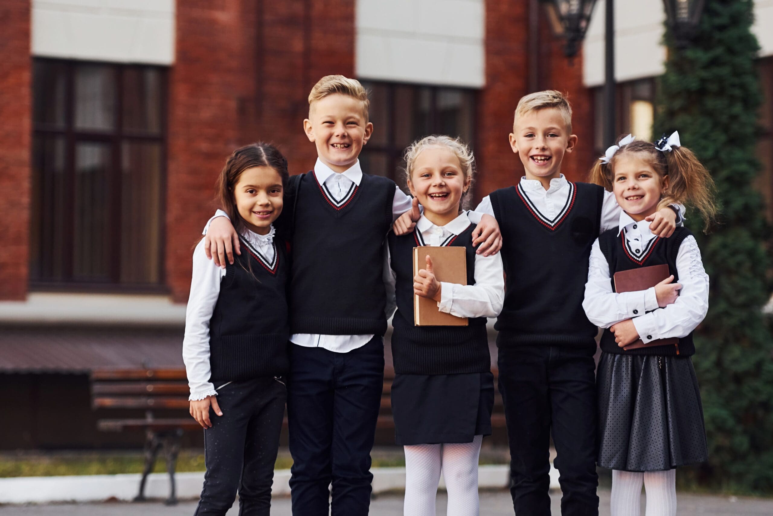 Five smiling children in school uniforms stand together outside a brick building, two holding books.