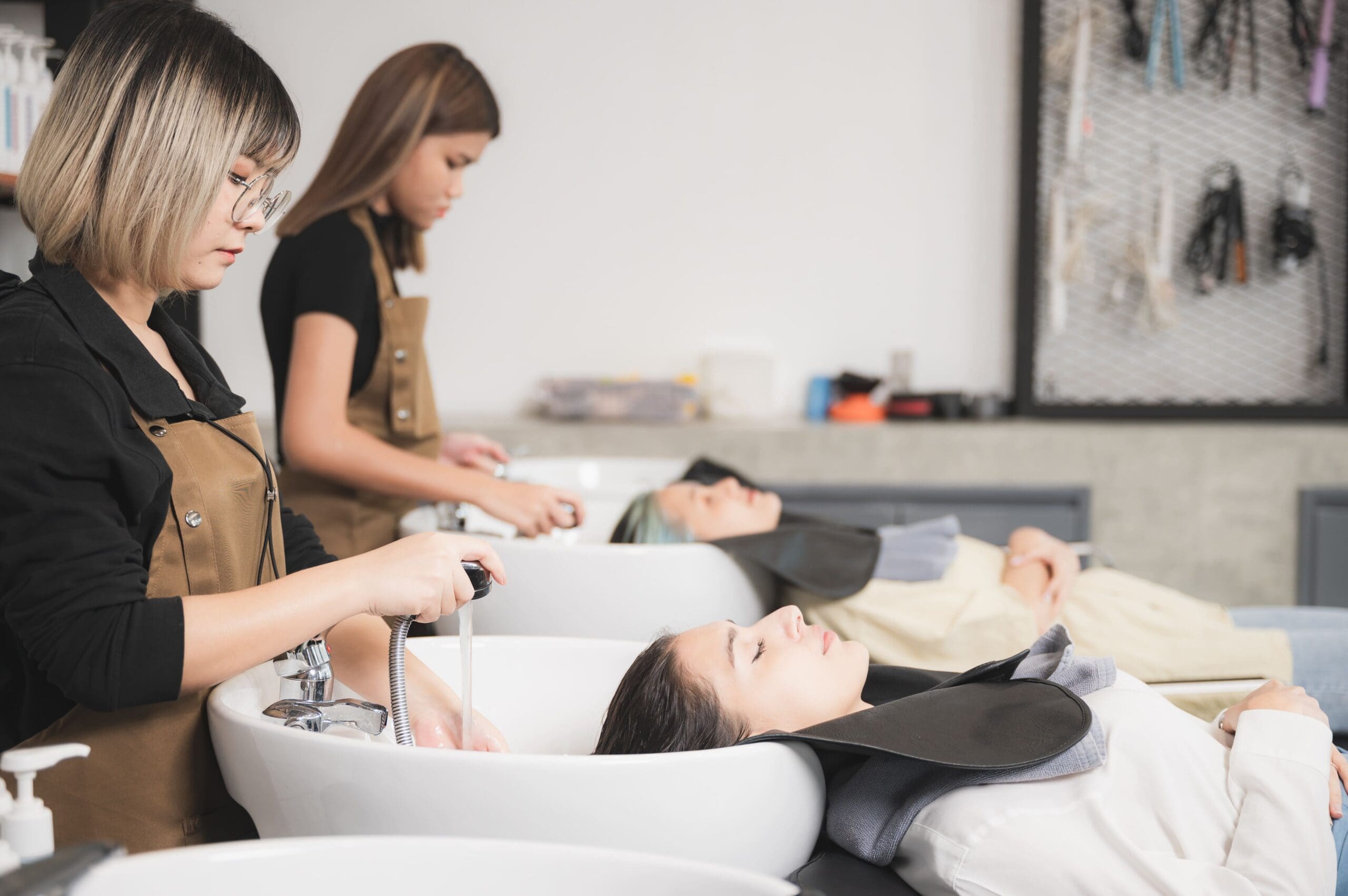 Two women get their hair washed at a salon, lying back in chairs, while stylists work over sinks.