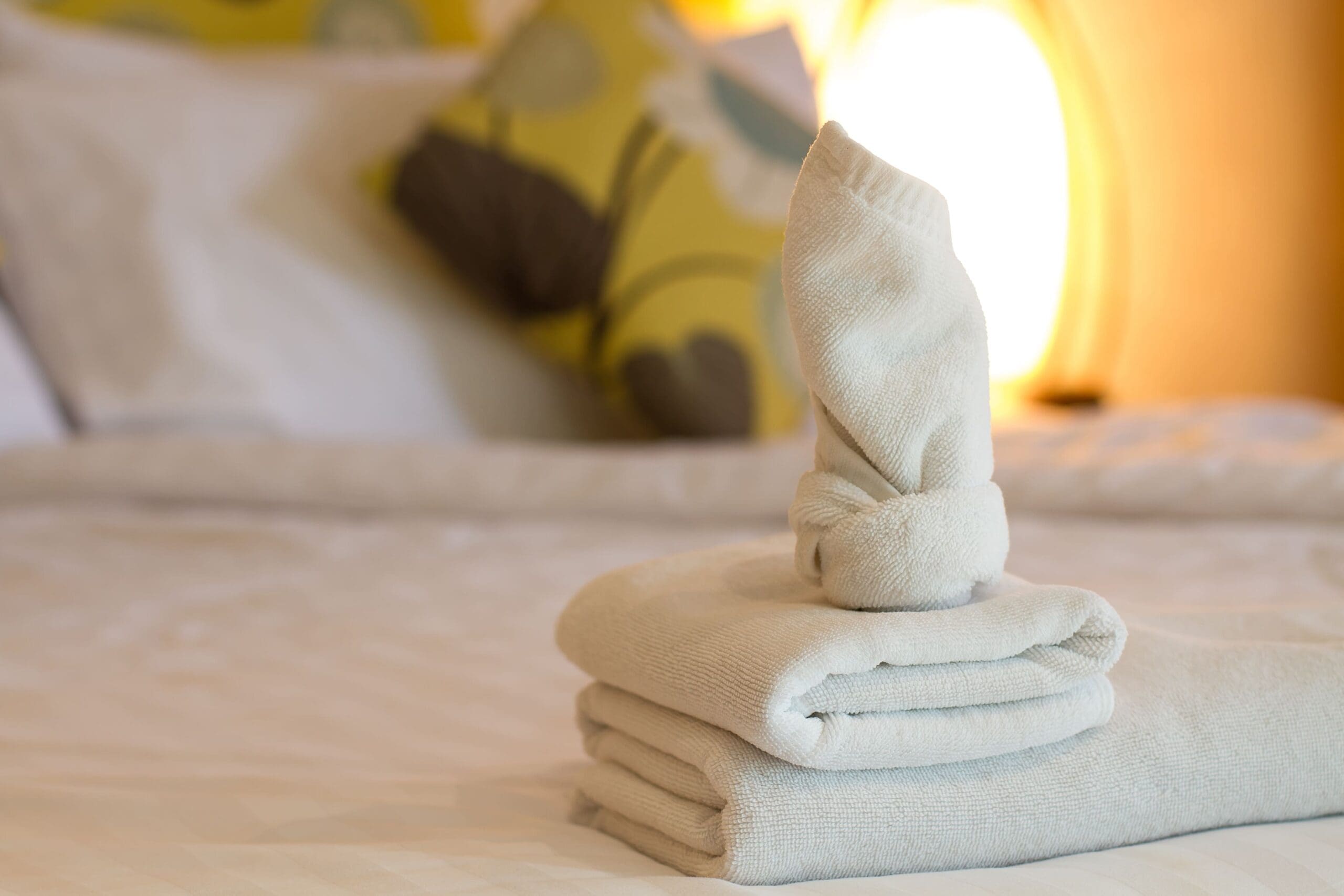 Two neatly folded towels with a towel shaped like a swan on top, set on a bed with patterned cushions and a lit lamp in the background.