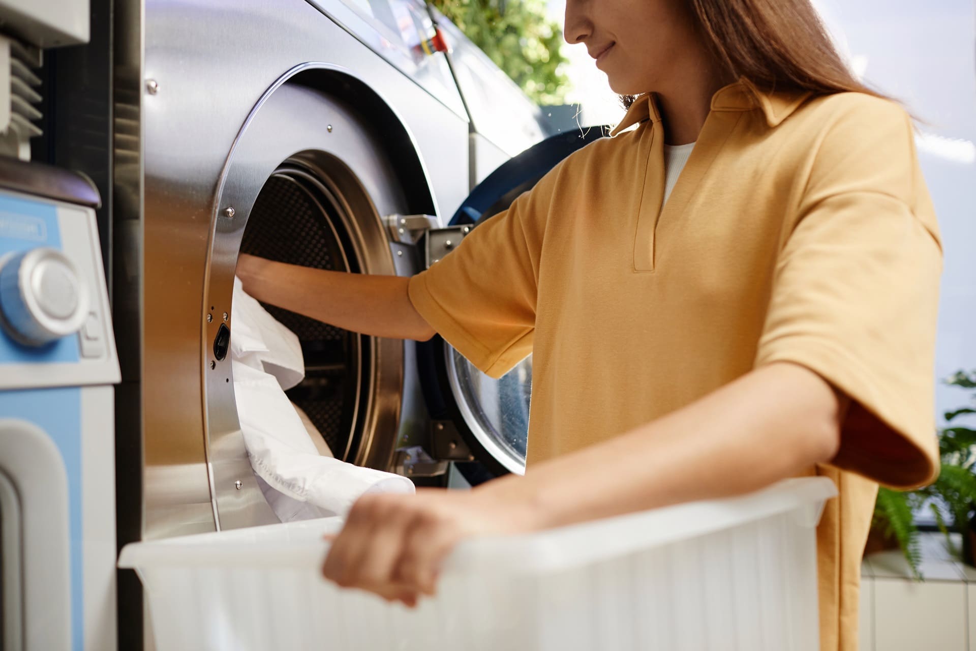 A person in a yellow shirt, having just found their perfect clothes cleaner near me, is unloading a white towel from a front-loading washing machine with a laundry basket conveniently placed nearby.