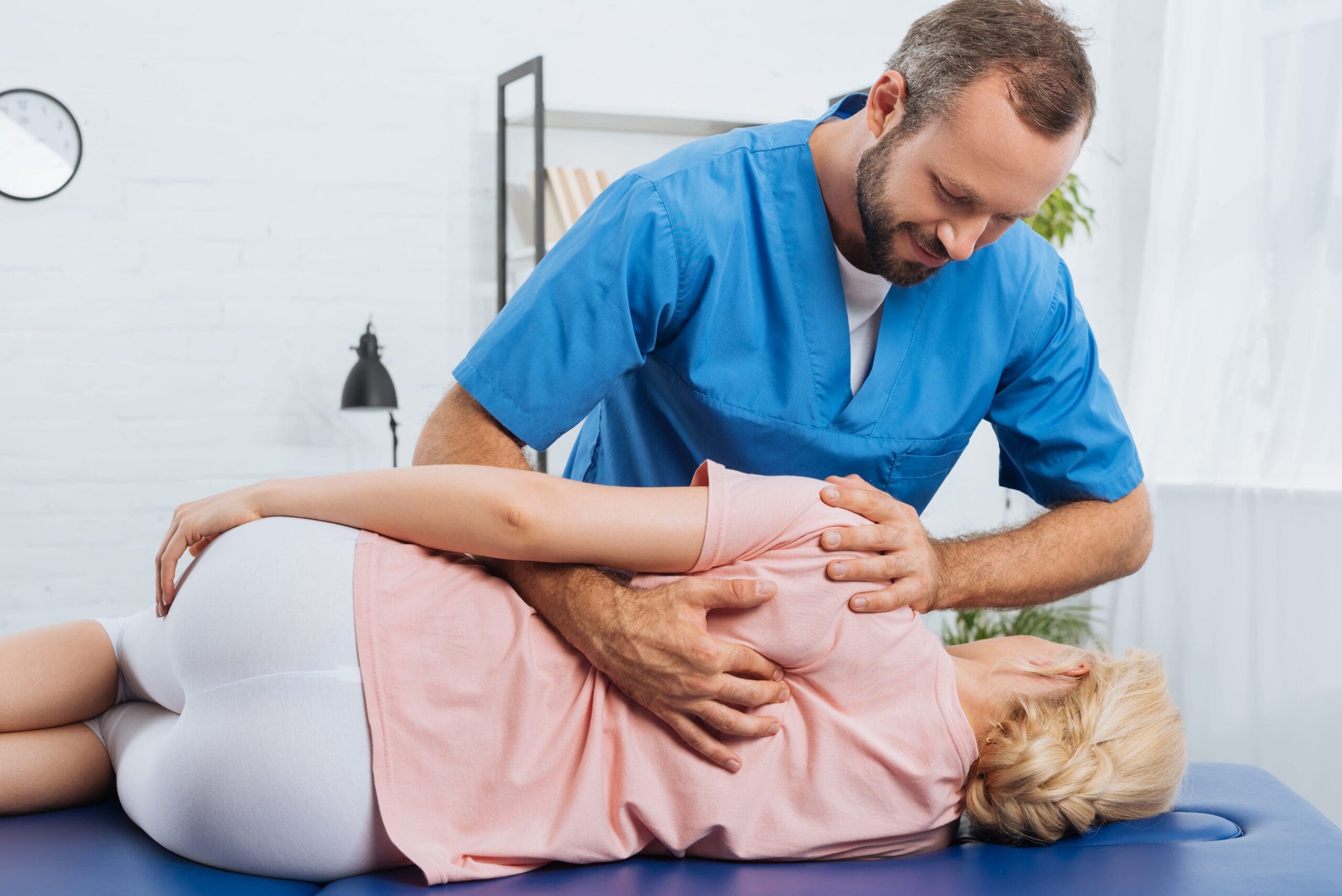 A chiropractor adjusts a woman's back as she lies on her side on a treatment table.