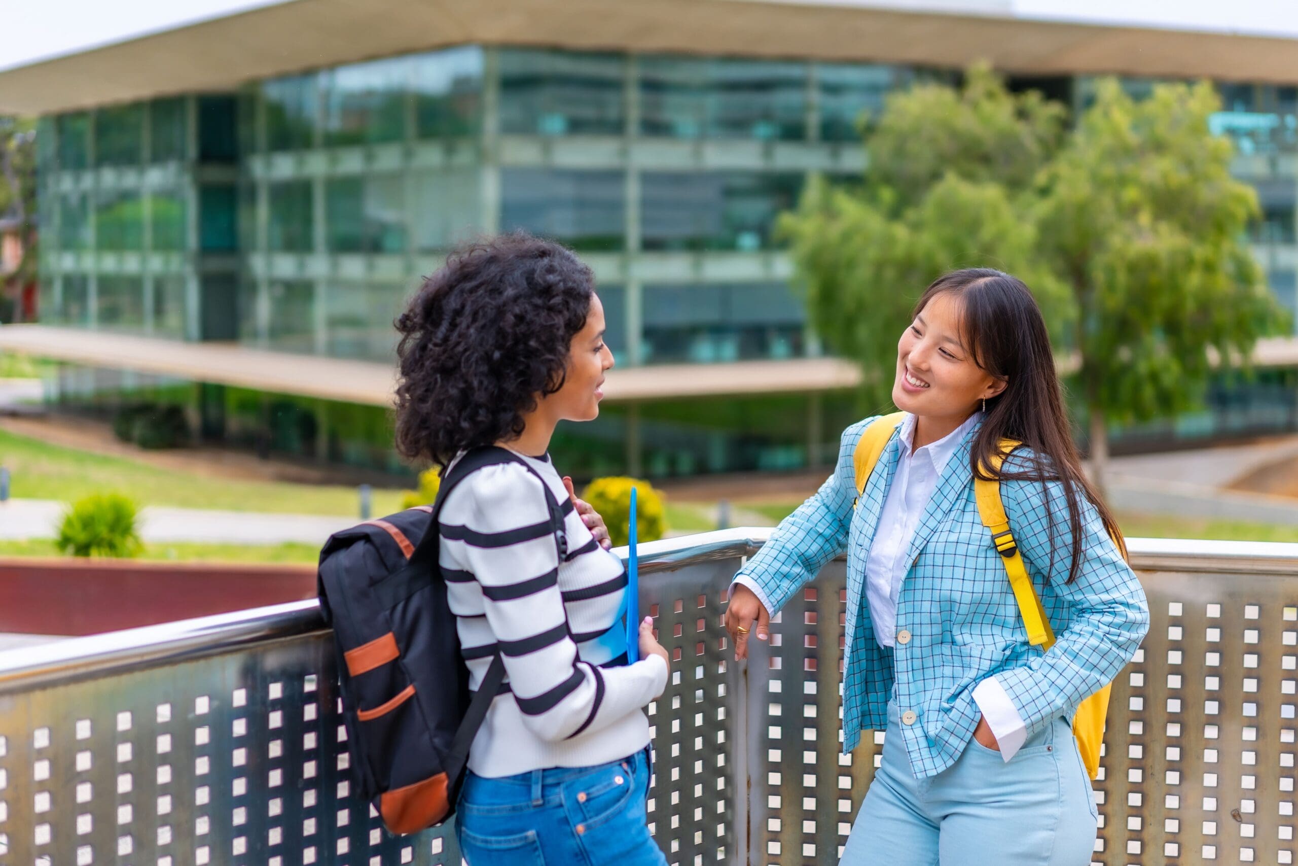 Two people with backpacks stand and chat on a metal railing in front of a modern glass building.