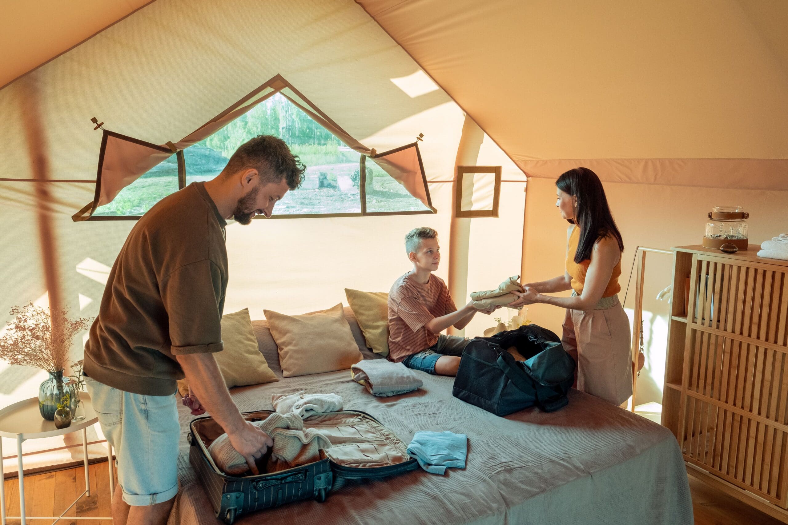 Three people are packing in a sunlit tent, organizing clothes into travel bags on a bed.