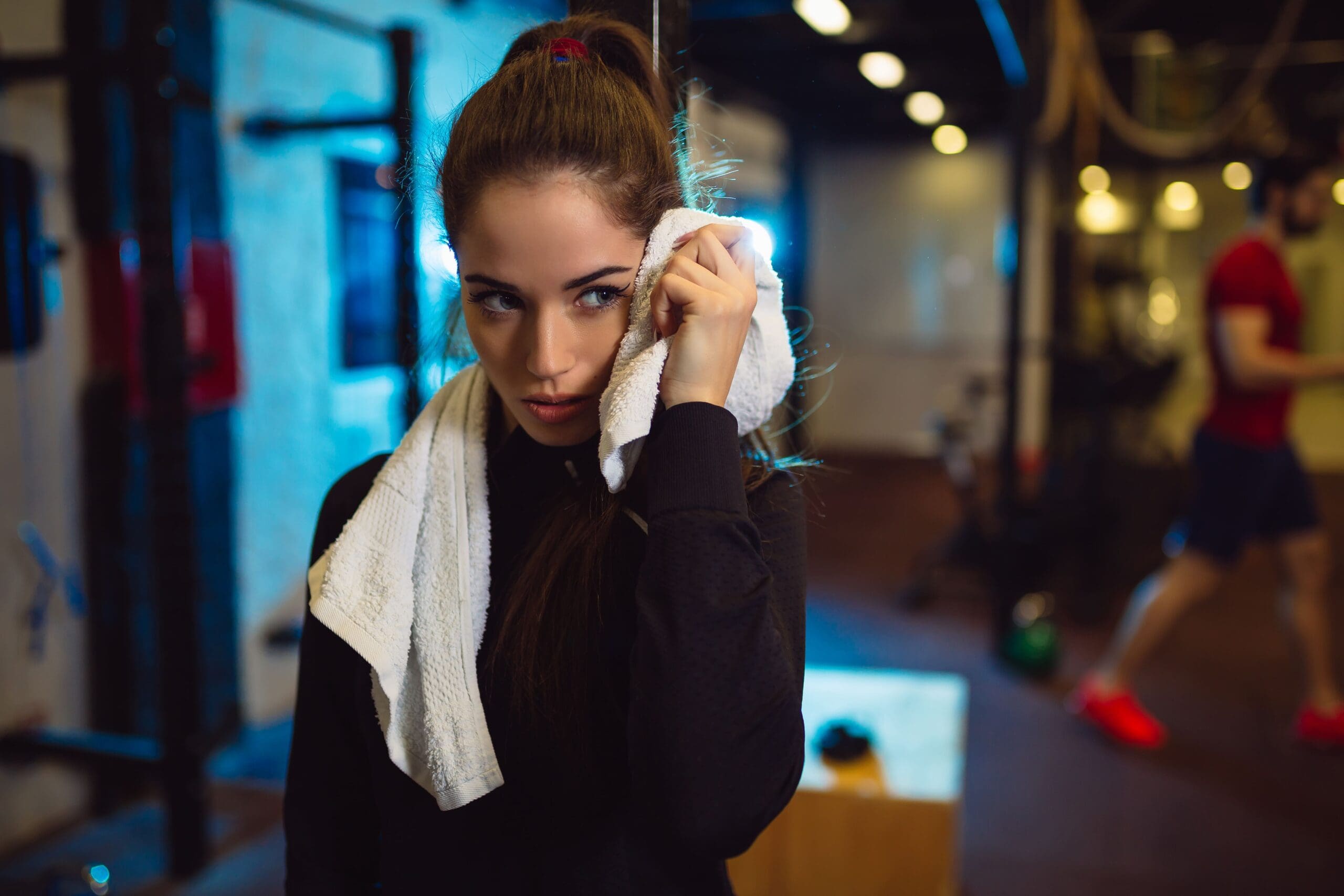 Woman in gym wiping her face with a towel, wearing a black top, with gym equipment and another person blurred in the background.