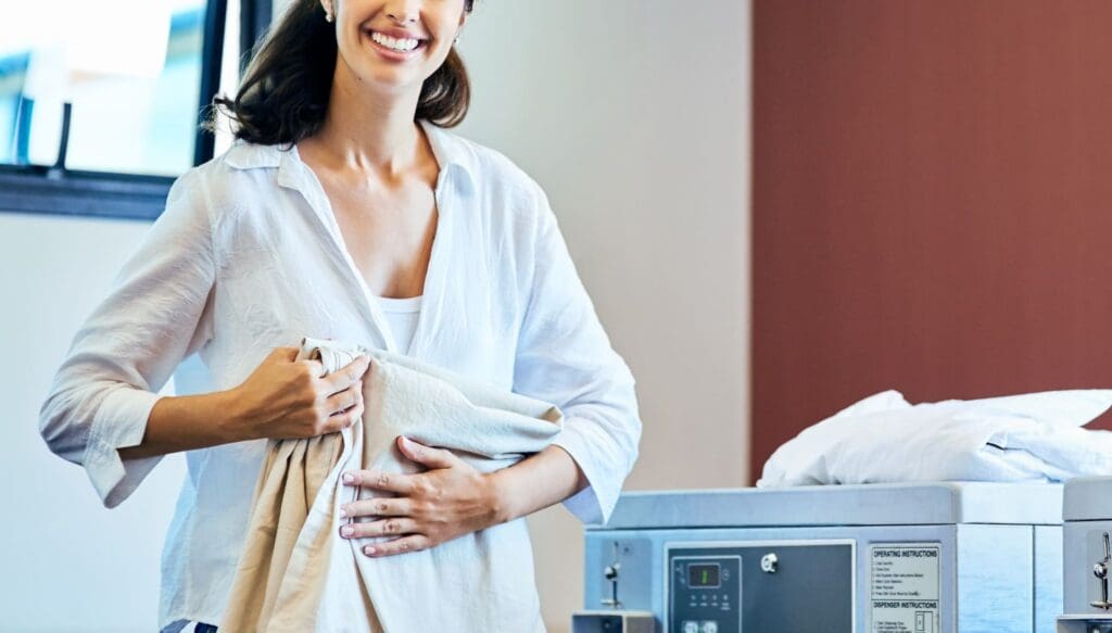 A person holding laundry stands next to a washing machine in a brightly lit room.