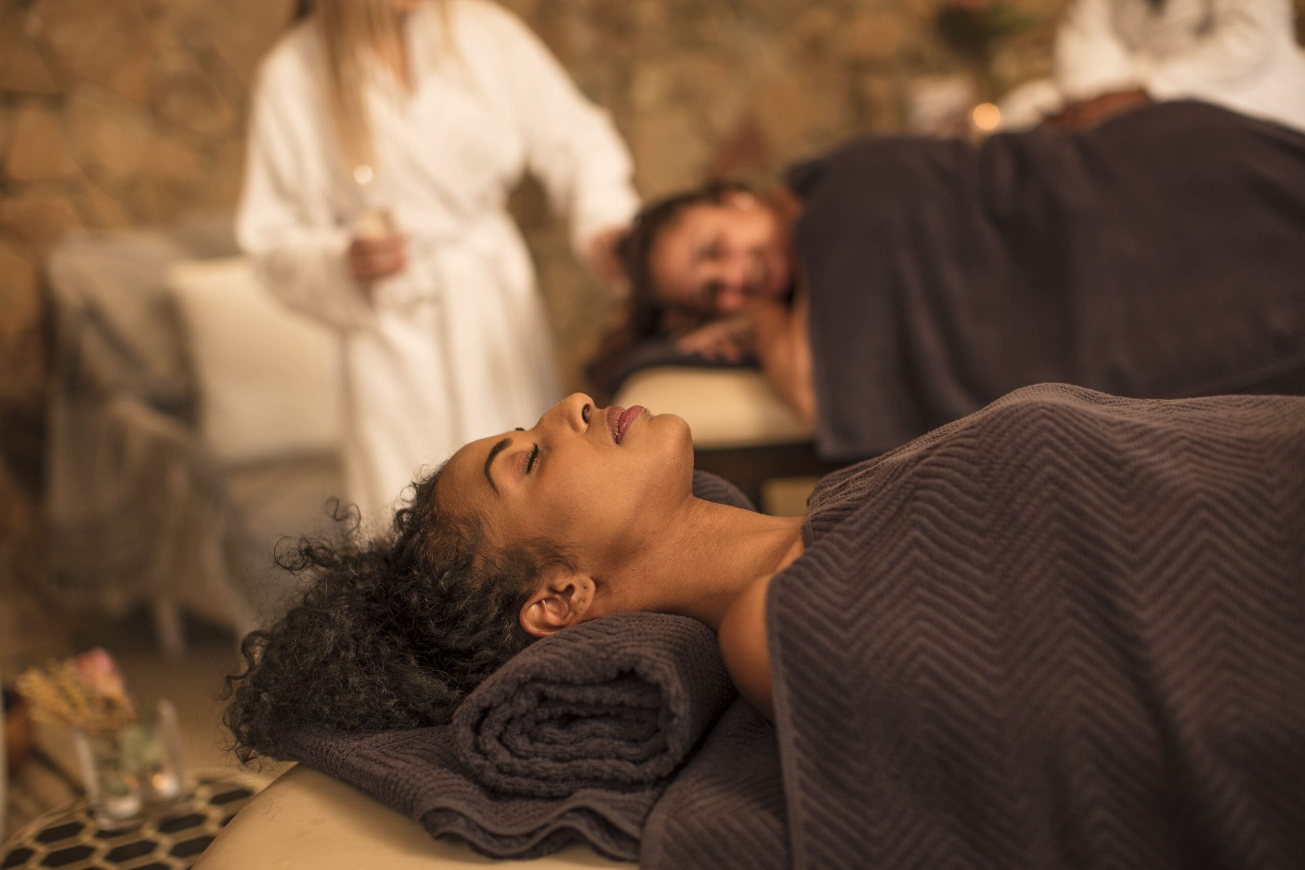 Two people are lying on massage tables, covered with dark towels. A person in a white robe stands nearby, holding a glass. The setting appears to be a spa or relaxation room.