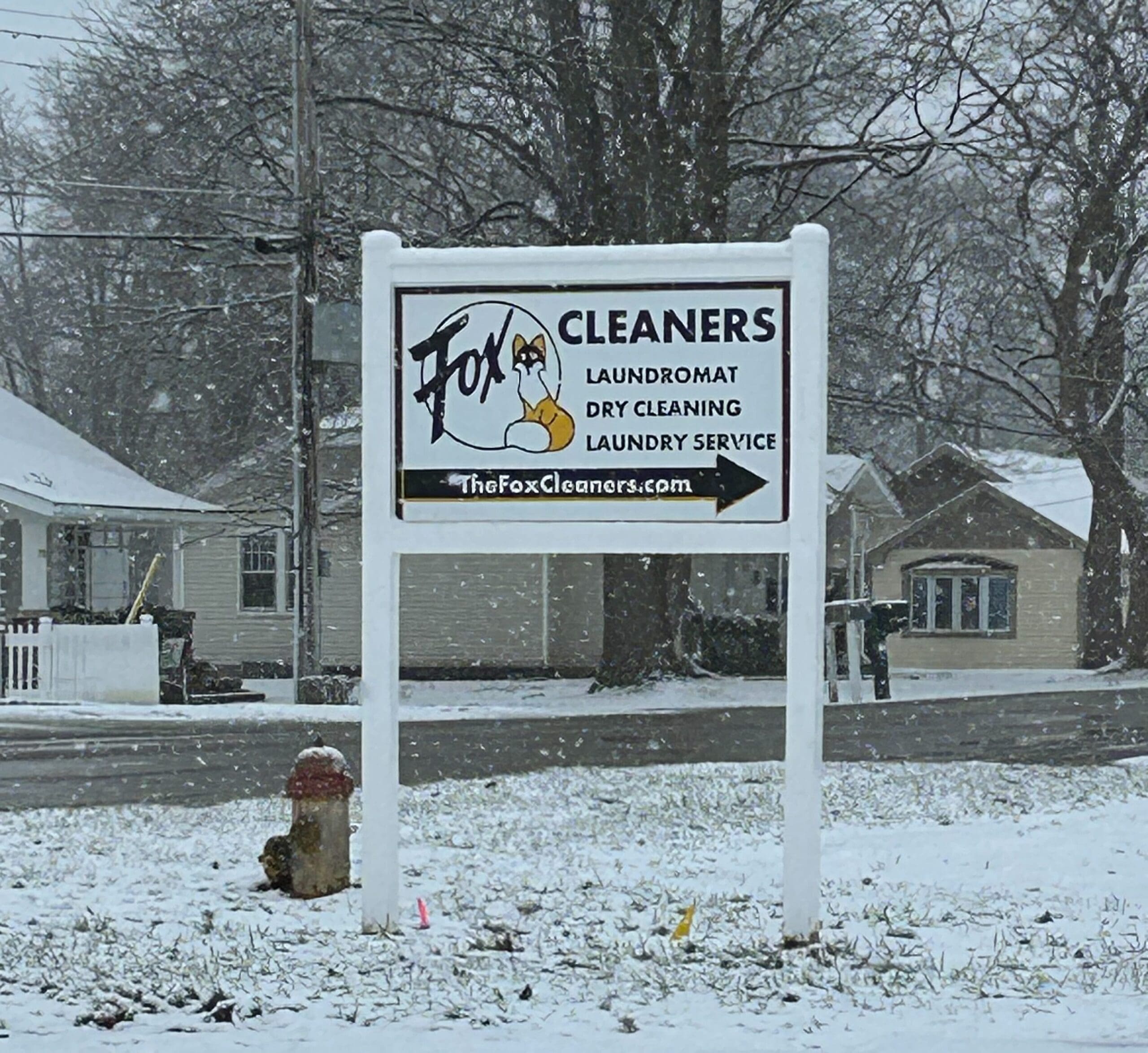 Sign for "The Fox Cleaners" in a snowy neighborhood, offering laundromat, dry cleaning, and laundry service, with an arrow pointing right.