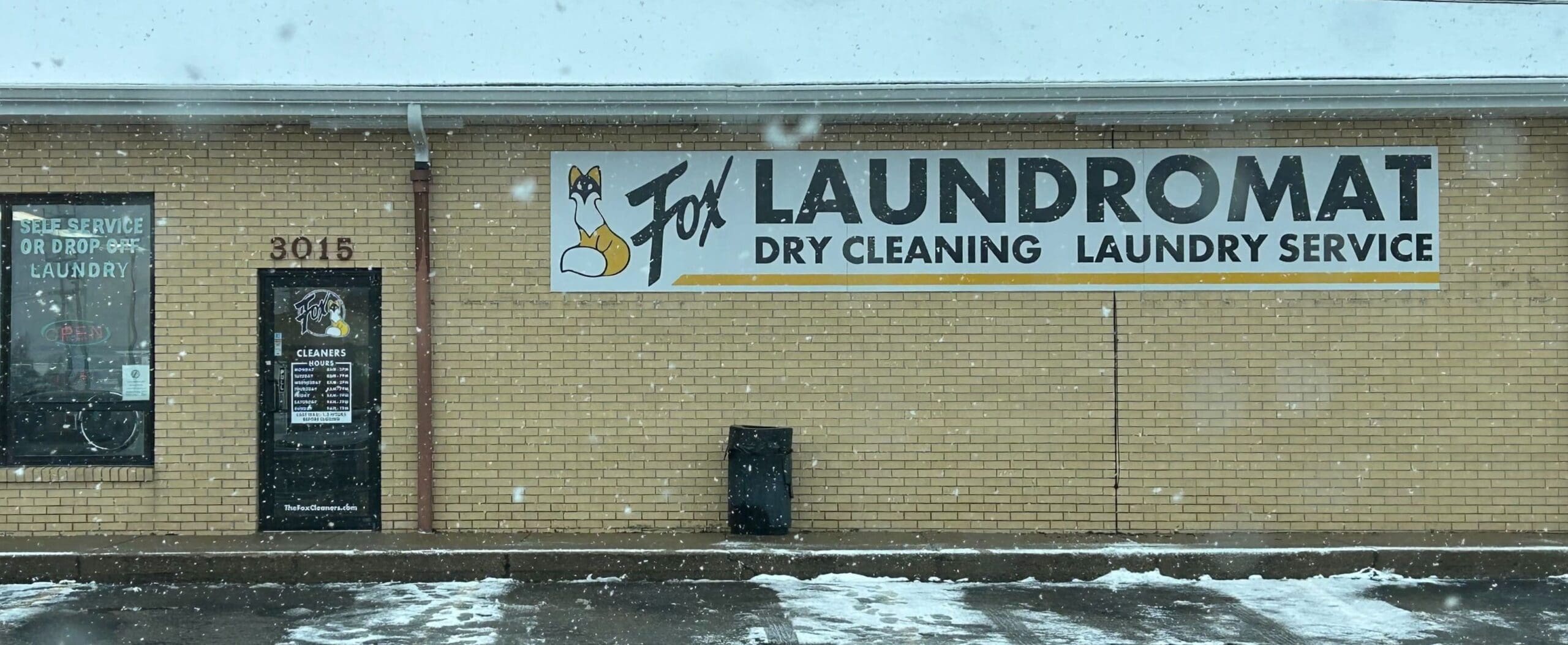 Laundromat with a sign for dry cleaning and laundry services on a brick building. Snow is visible on the ground.