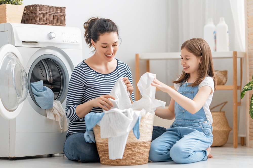 Woman and young girl sitting on the floor doing laundry together, smiling, with a front-load washing machine and a laundry basket filled with clothes nearby.